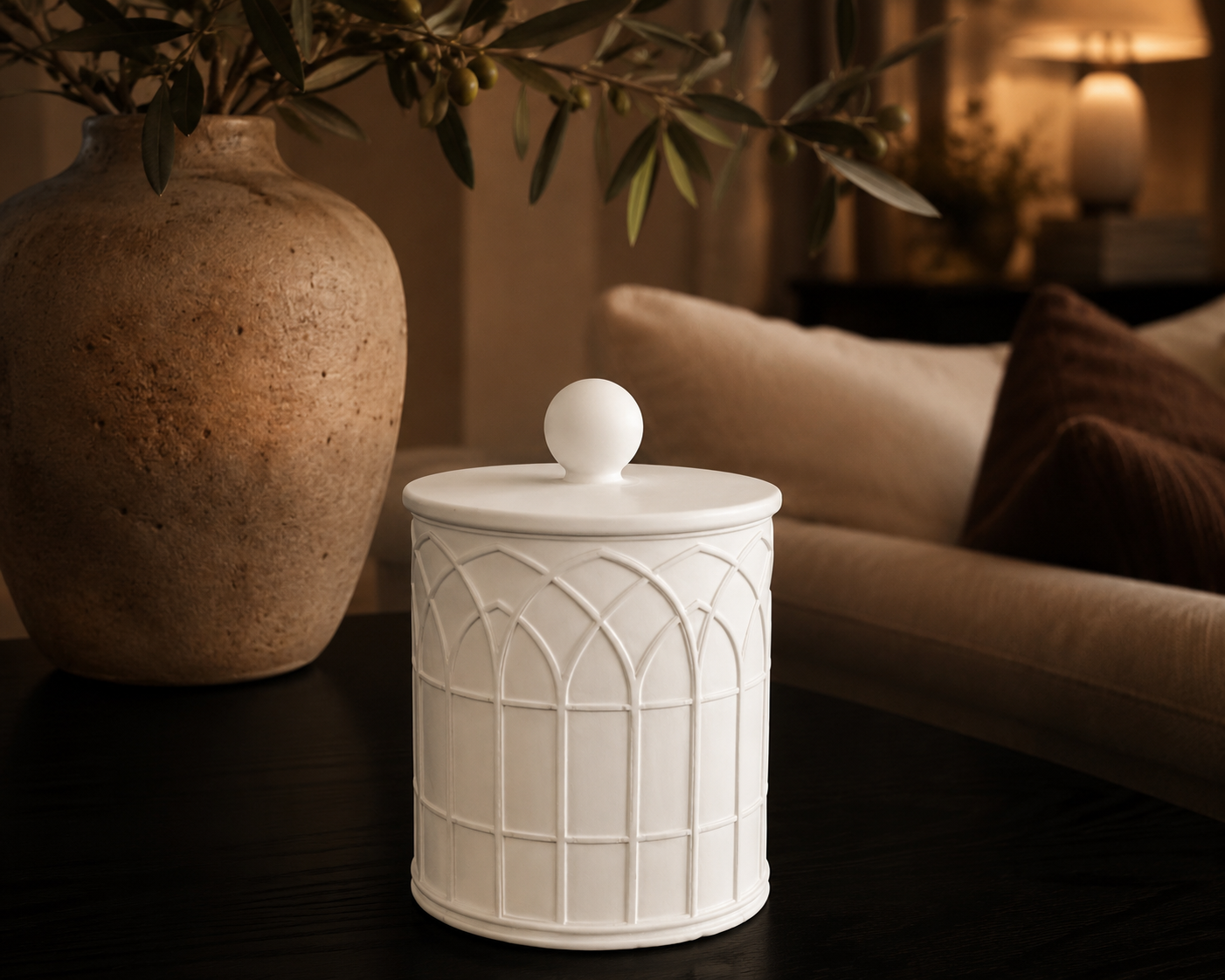 Arcade Stone Vessel styled with its ball-topped lid on a dark wooden table beside an olive branch in a terracotta vase, in a warm lamp-lit living room